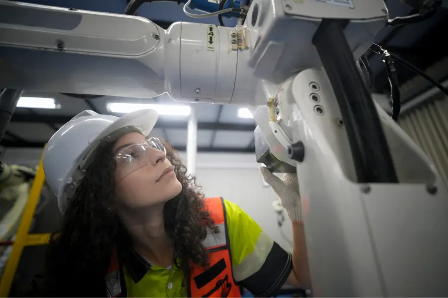Engineer in a hard hat, safety glasses, and high-visibility vest inspecting a white industrial robotic arm on a factory floor.